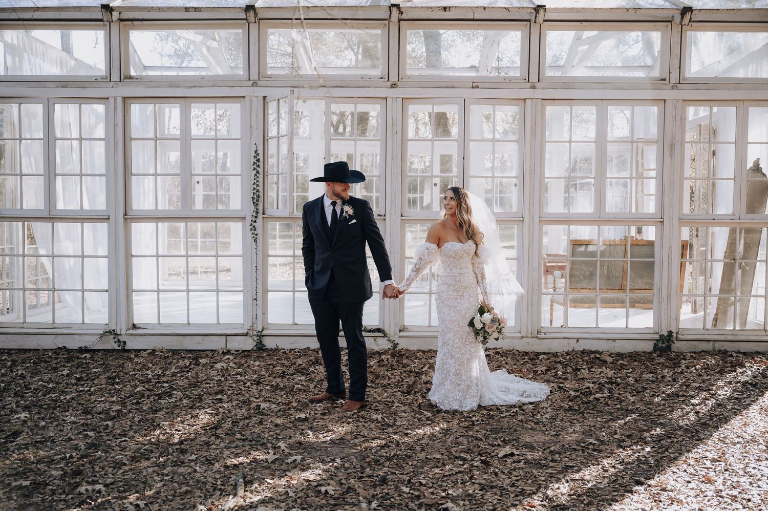 Stylish wedding couple poses in front of greenhouse windows in modern attire.