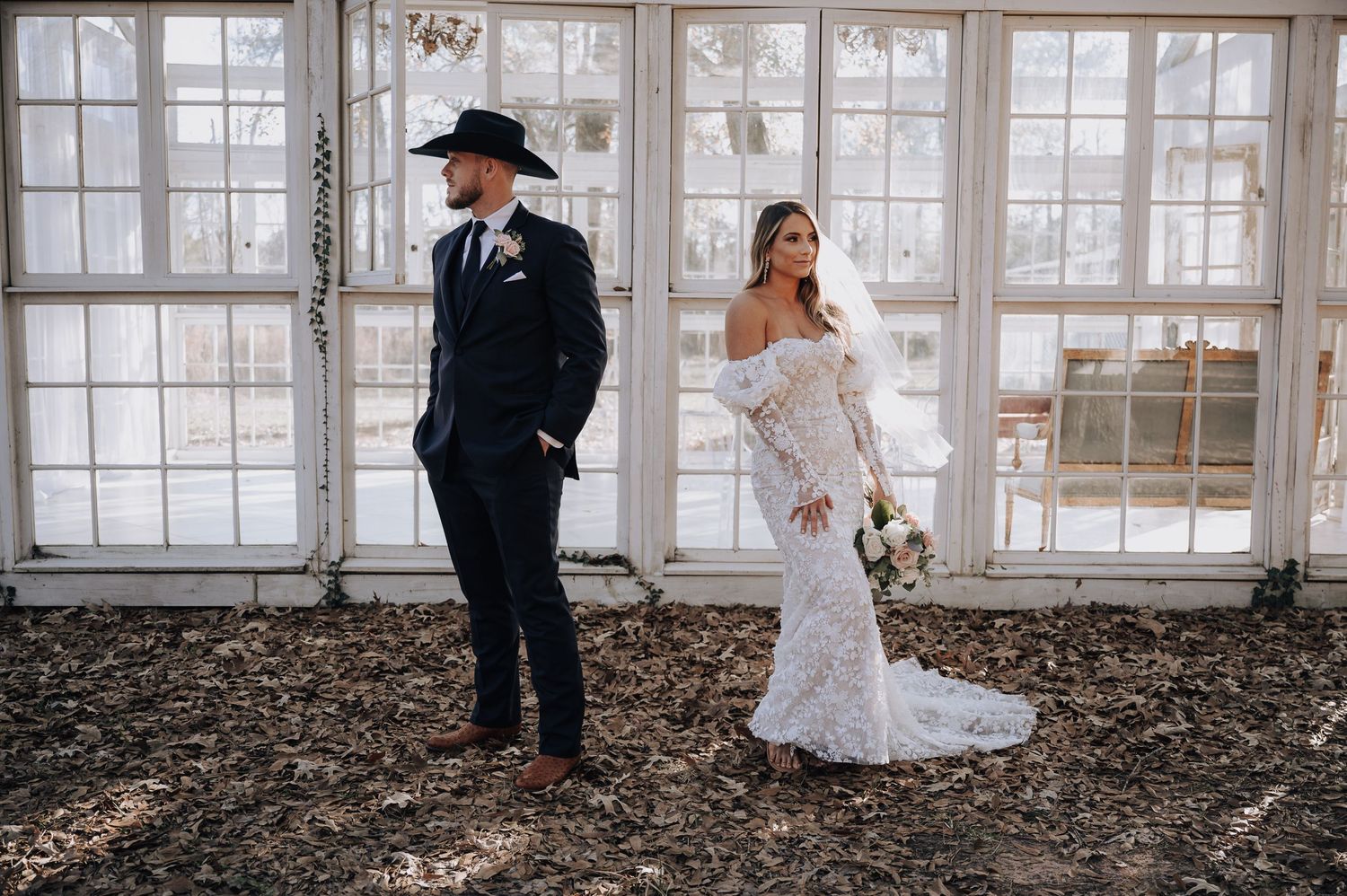 Artistic wedding portrait of couple in formal attire standing apart in greenhouse setting.