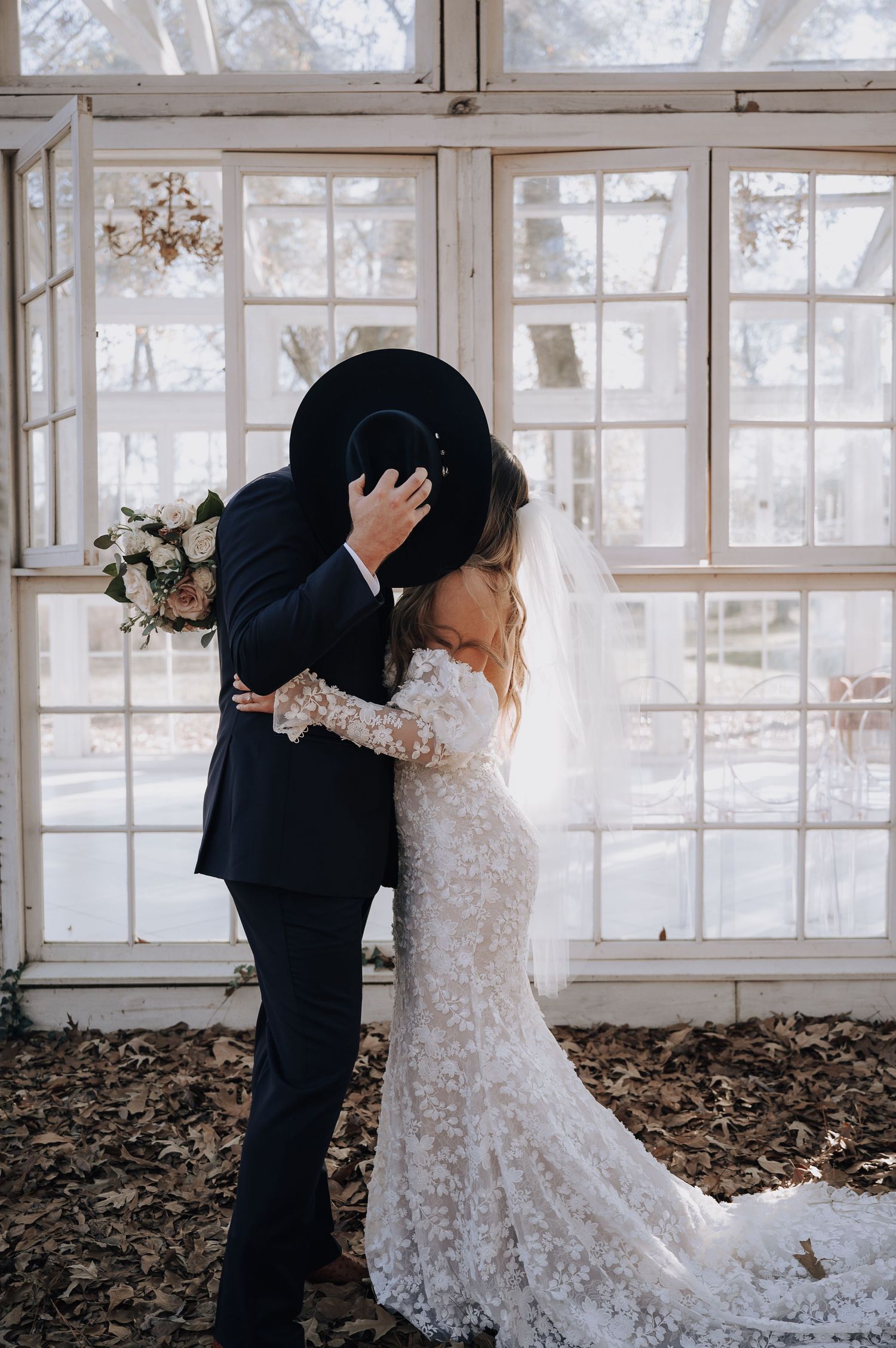 A romantic wedding photo of a couple embracing in front of sunlit windows, with the bride wearing a lace gown.