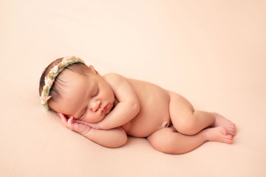 Peaceful sleeping newborn baby wearing floral headband against peach background.