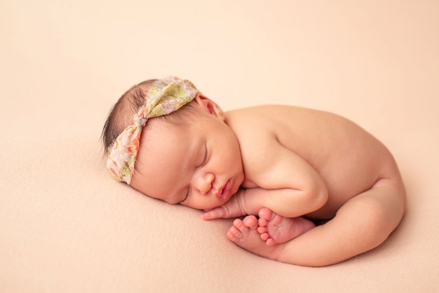 Sleeping newborn baby poses with floral headband on peachy pink backdrop.