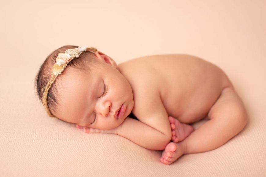 Sleeping newborn baby wearing delicate floral headband on soft peach background.