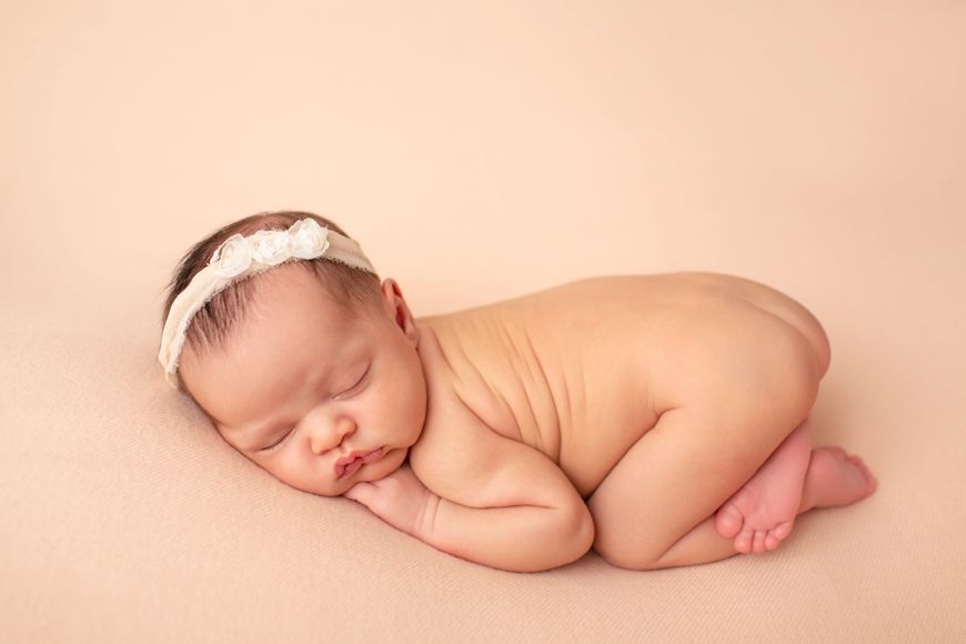 Sleeping newborn baby curled up with headband on peachy backdrop.