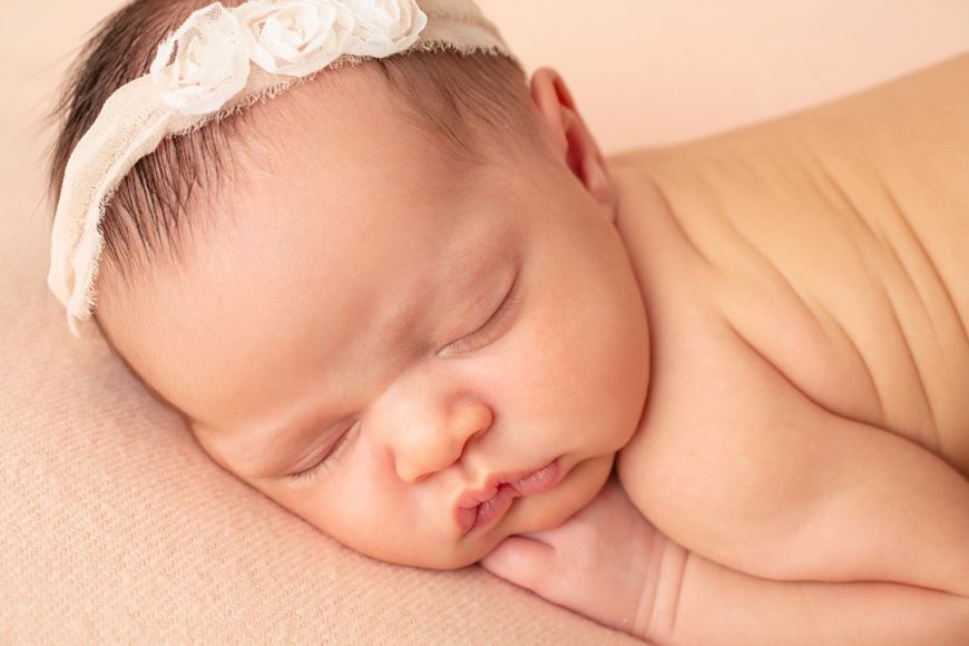 Sleeping newborn wearing delicate white lace headband on pink background.