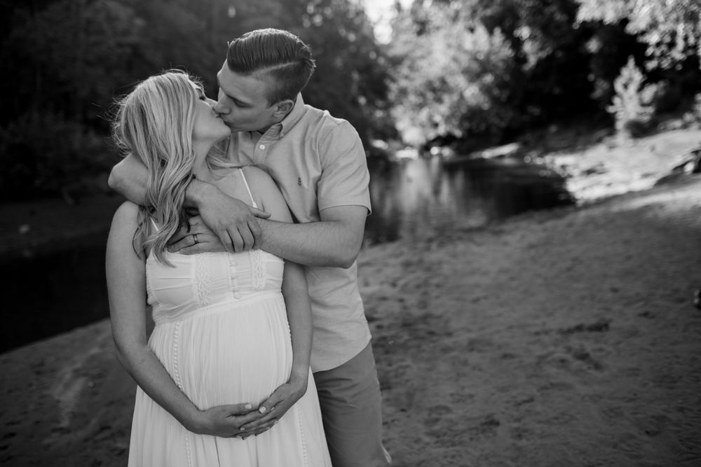 Black and white maternity photo of a couple sharing a kiss by a peaceful riverside.