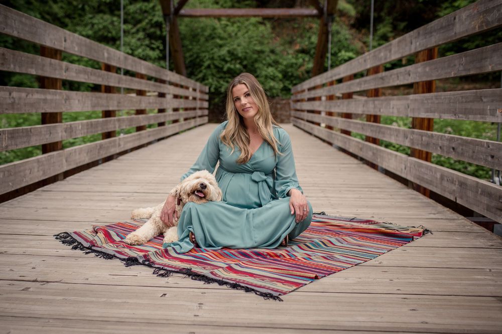 A pregnant woman in a teal dress sits on a colorful rug on a wooden bridge with her dog.