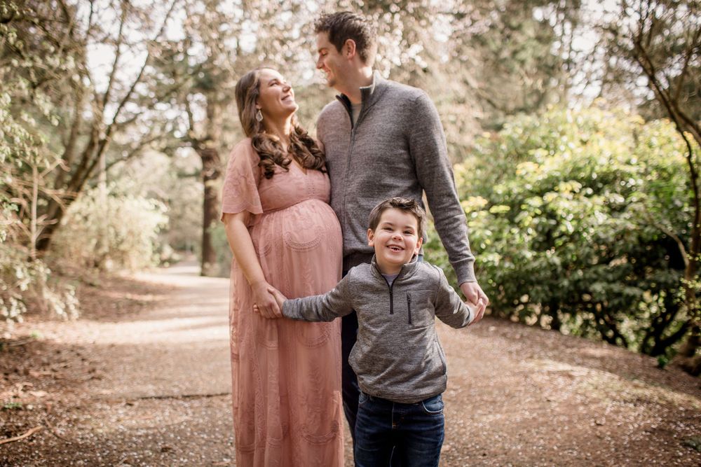 A pregnant woman in a pink dress stands with her partner and young son on a tree-lined path.