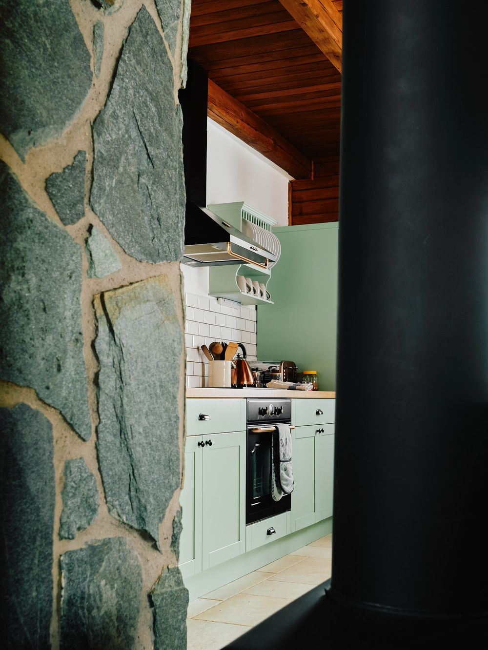 Modern kitchen with mint green cabinets viewed through stone archway with wooden ceiling.