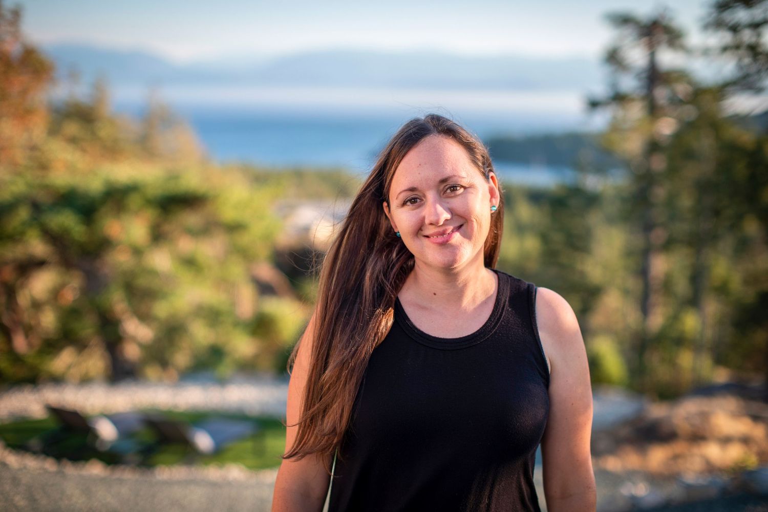 Smiling woman with long brown hair standing outdoors with ocean and trees in background.