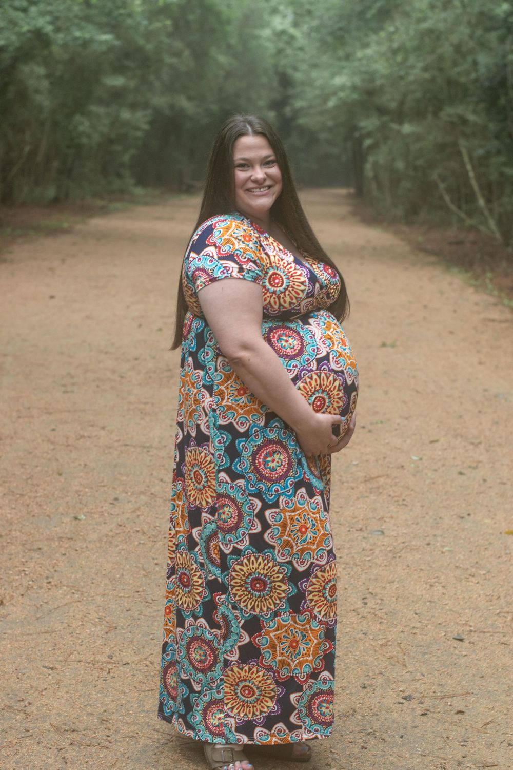A pregnant woman in a vibrant floral maxi dress standing on a forest path.