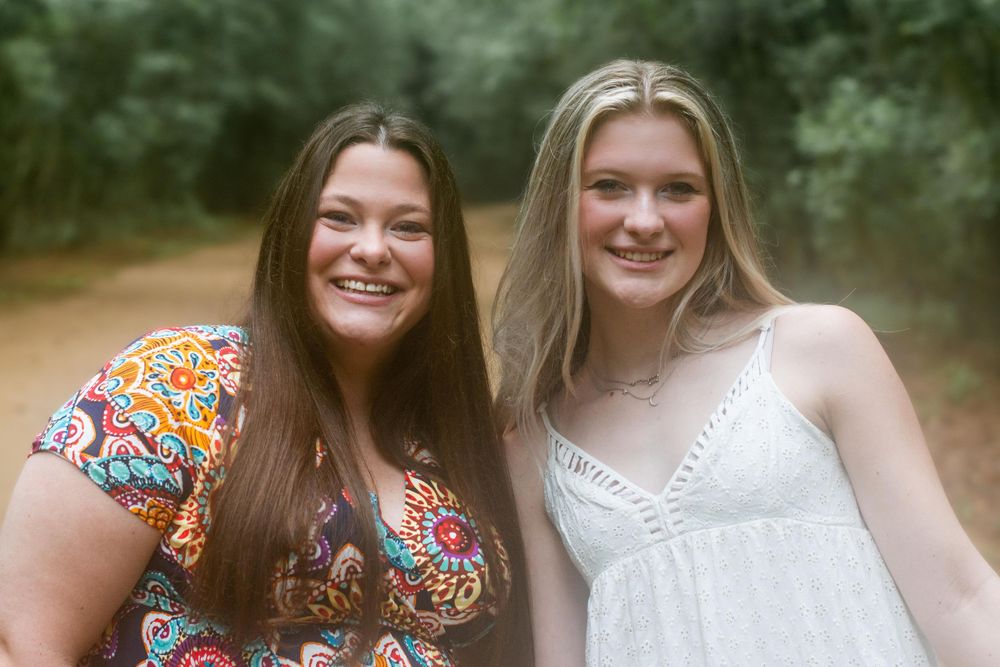 Two friends pose together on a wooded trail wearing contrasting summer dresses.