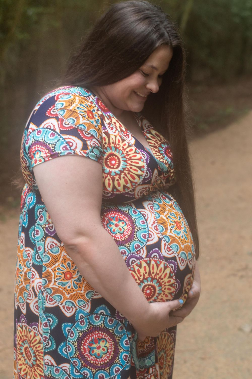 A pregnant woman in patterned dress smiles while touching her baby bump.