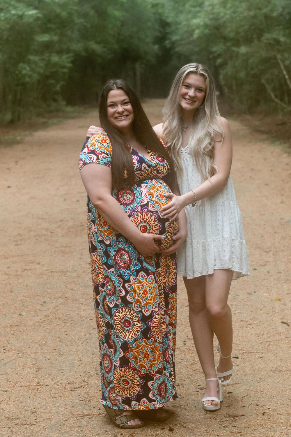 Two friends share a tender moment during maternity photoshoot on forest trail.