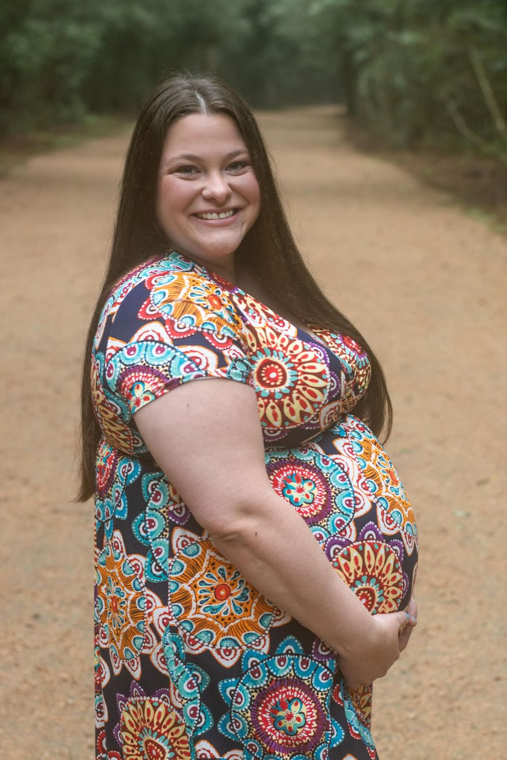 A smiling pregnant woman poses on forest path wearing floral maxi dress.