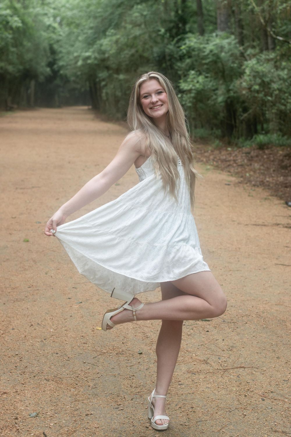 A woman in white dress twirls playfully on a forest path.