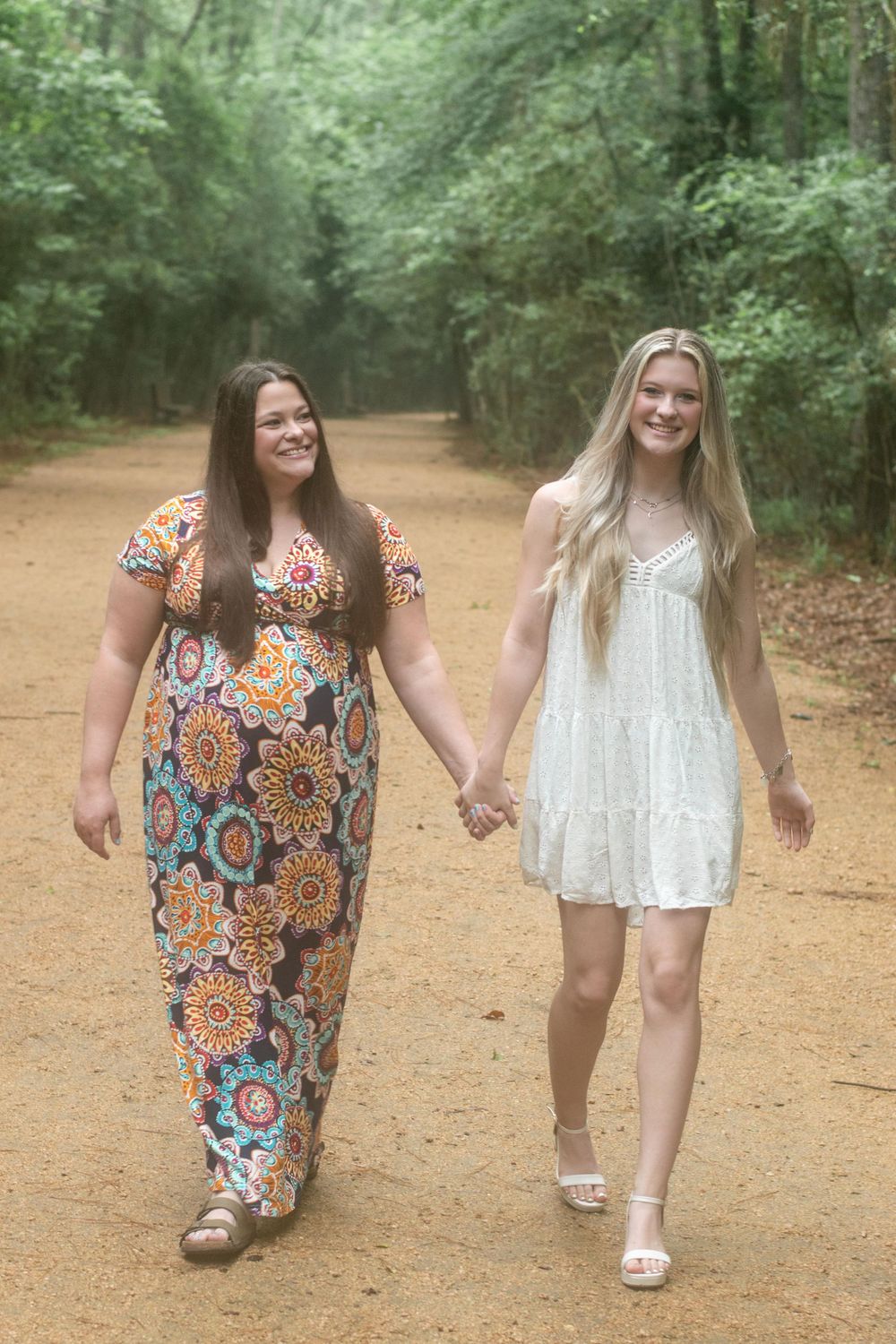 Two women walking together on a dirt path through lush green forest.