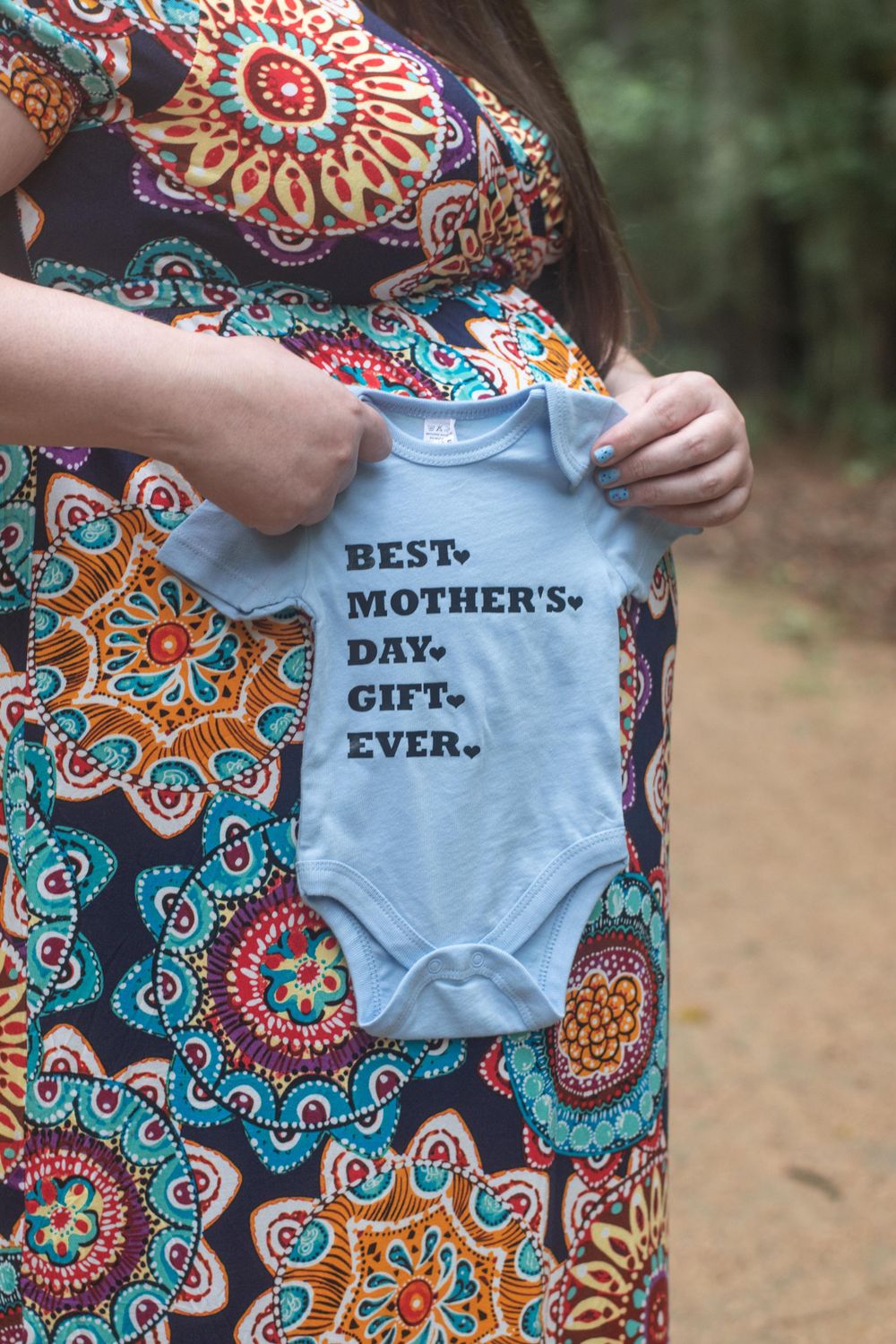 A baby onesie with 'Best Mother's Day Gift Ever' text held against a colorful floral print maternity dress.