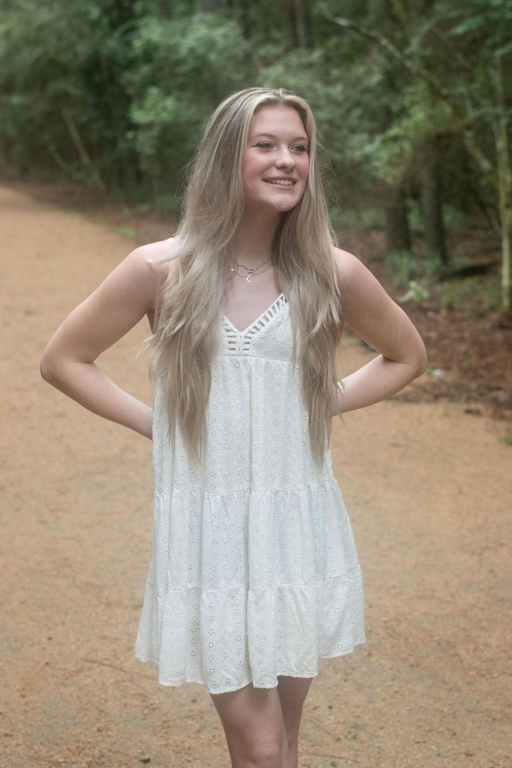 A woman in a white summer dress poses on a dirt path surrounded by trees.