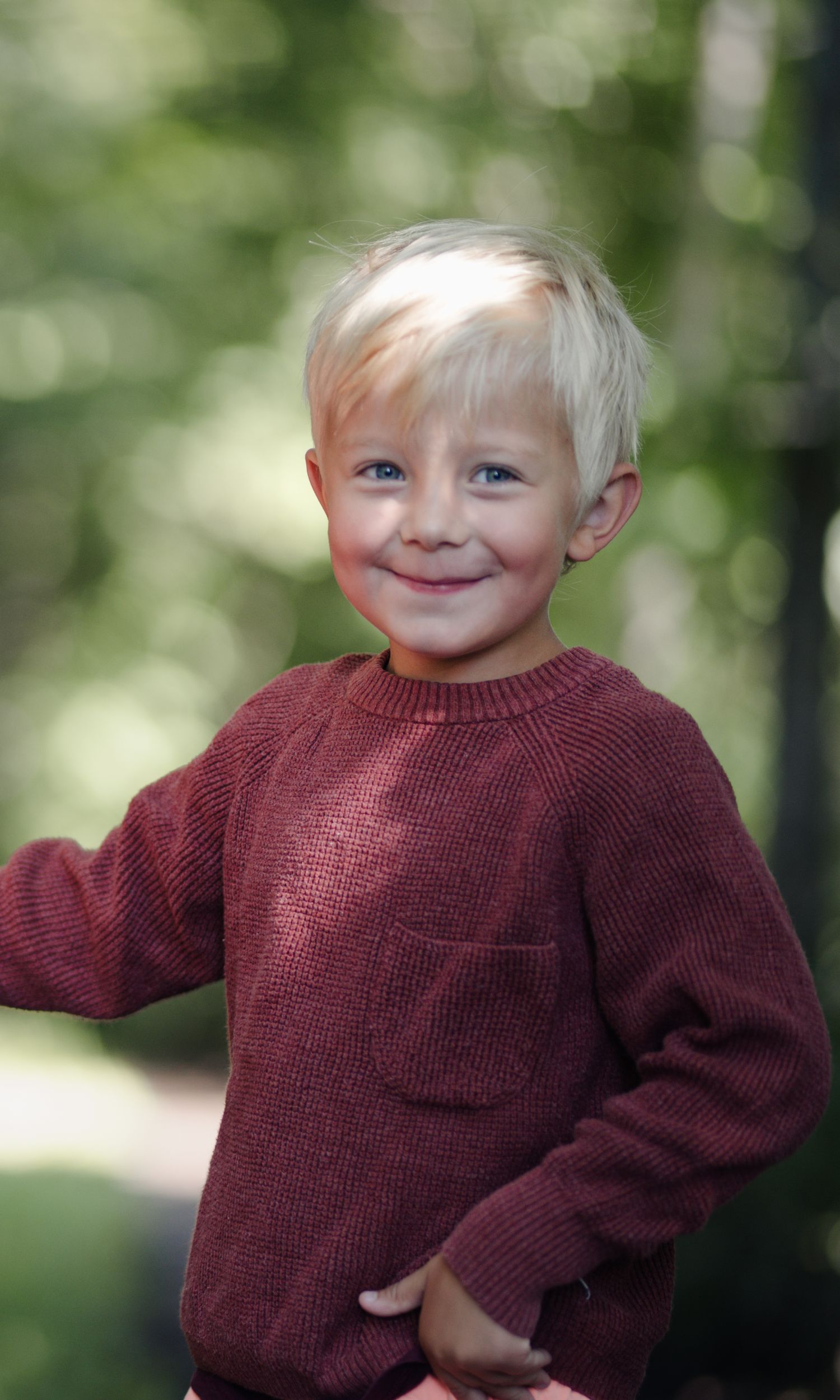 Child in burgundy sweater smiles warmly at camera in outdoor portrait.
