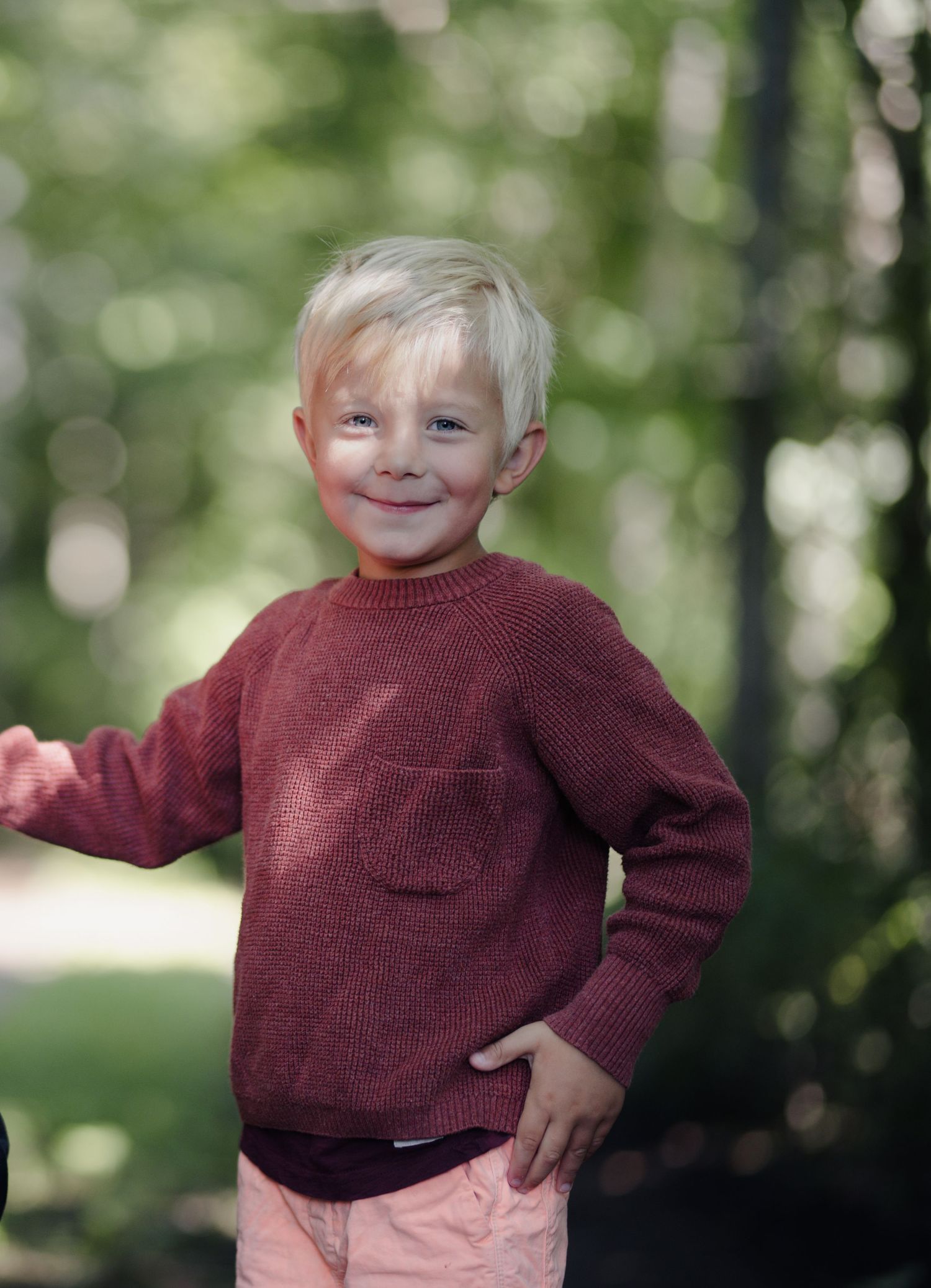Child poses playfully in burgundy sweater against blurred forest backdrop.