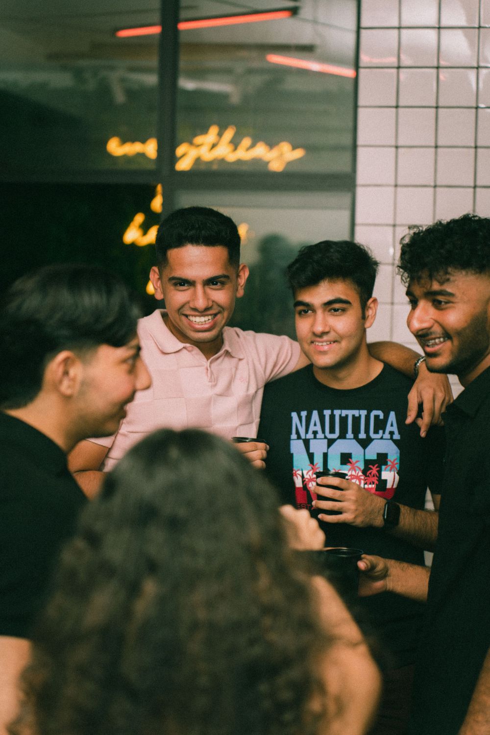 Group of friends sharing laughs and drinks under neon lighting at a social gathering.