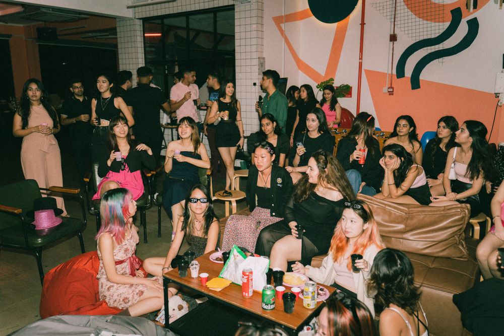 Large group of party guests socializing in a decorated indoor space with pink accents.