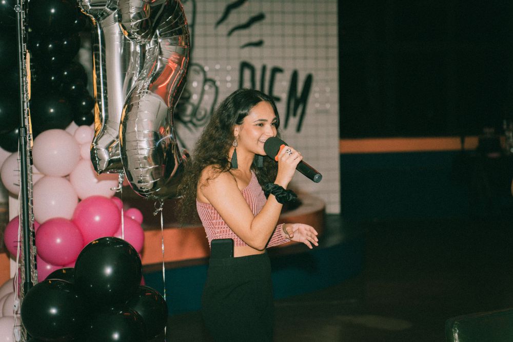 Person holding a microphone performing at an event decorated with pink and silver balloons.