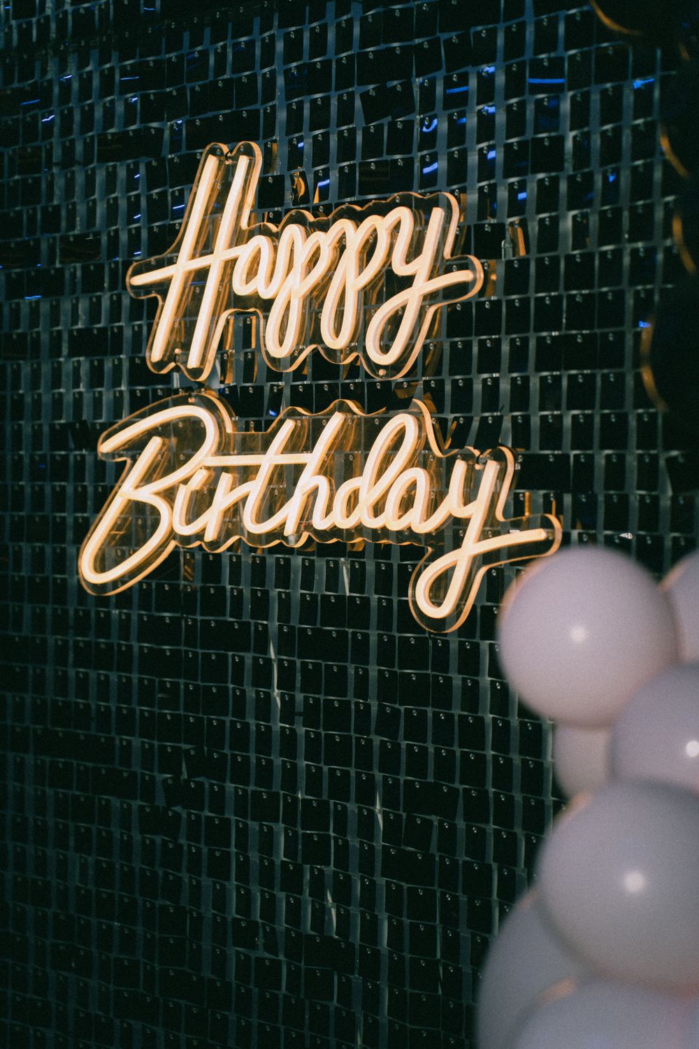 Glowing neon 'Happy Birthday' sign mounted on dark textured wall with white balloons.