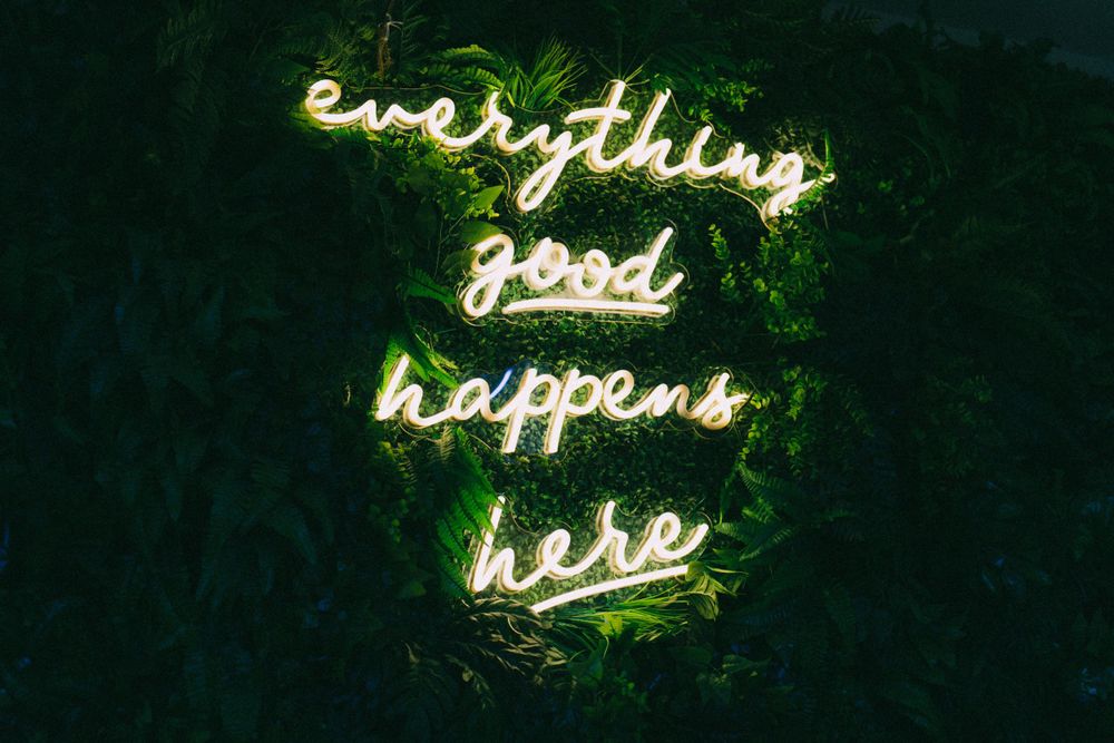 Neon green sign reading 'everything good happens here' glowing against dark background.