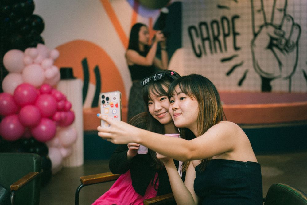 Two friends taking a selfie at a party with pink balloons and decorative wall art in the background.