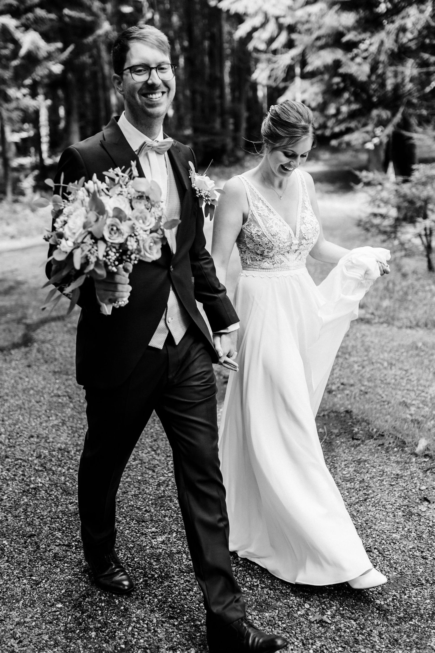 A joyful wedding couple walks through a wooded area carrying a beautiful floral bouquet.