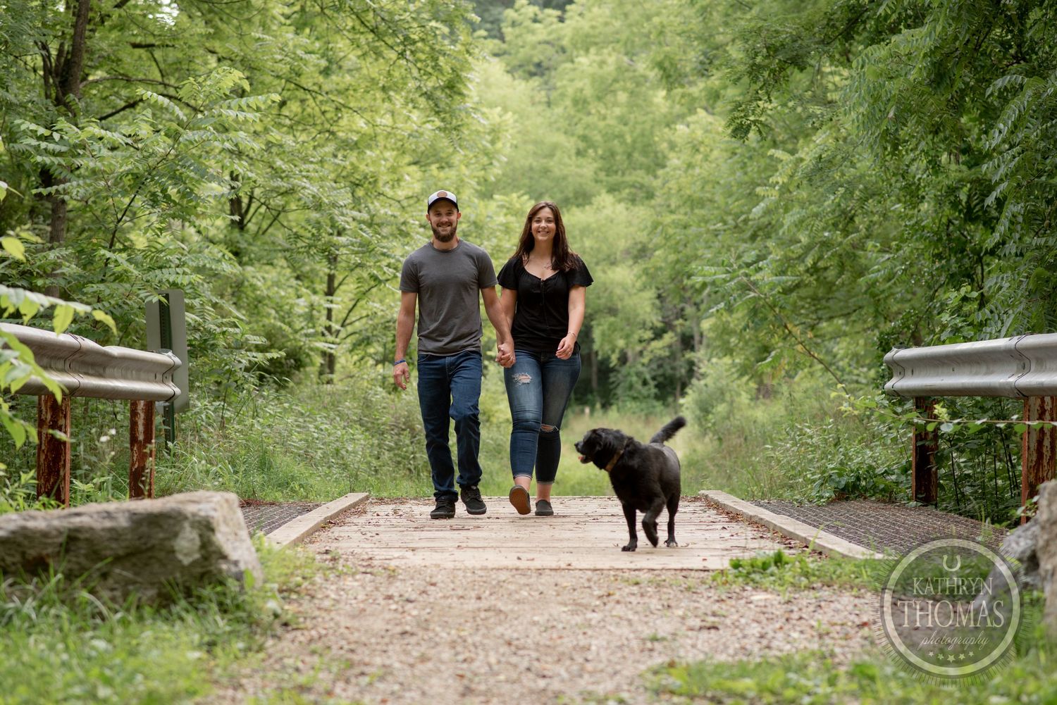 A couple walks their black dog along a tree-lined path with guard rails.