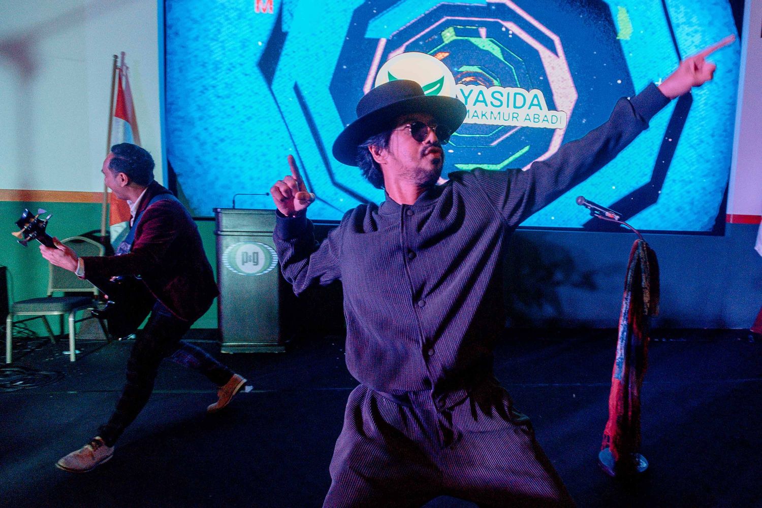 Performer in dark clothing and hat dances against a vibrant blue illuminated backdrop.