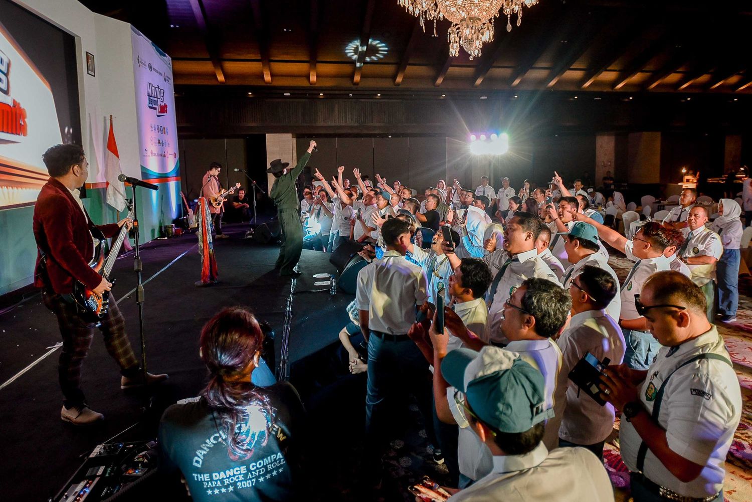 Wide concert shot showing performers on stage with enthusiastic crowd raising hands.