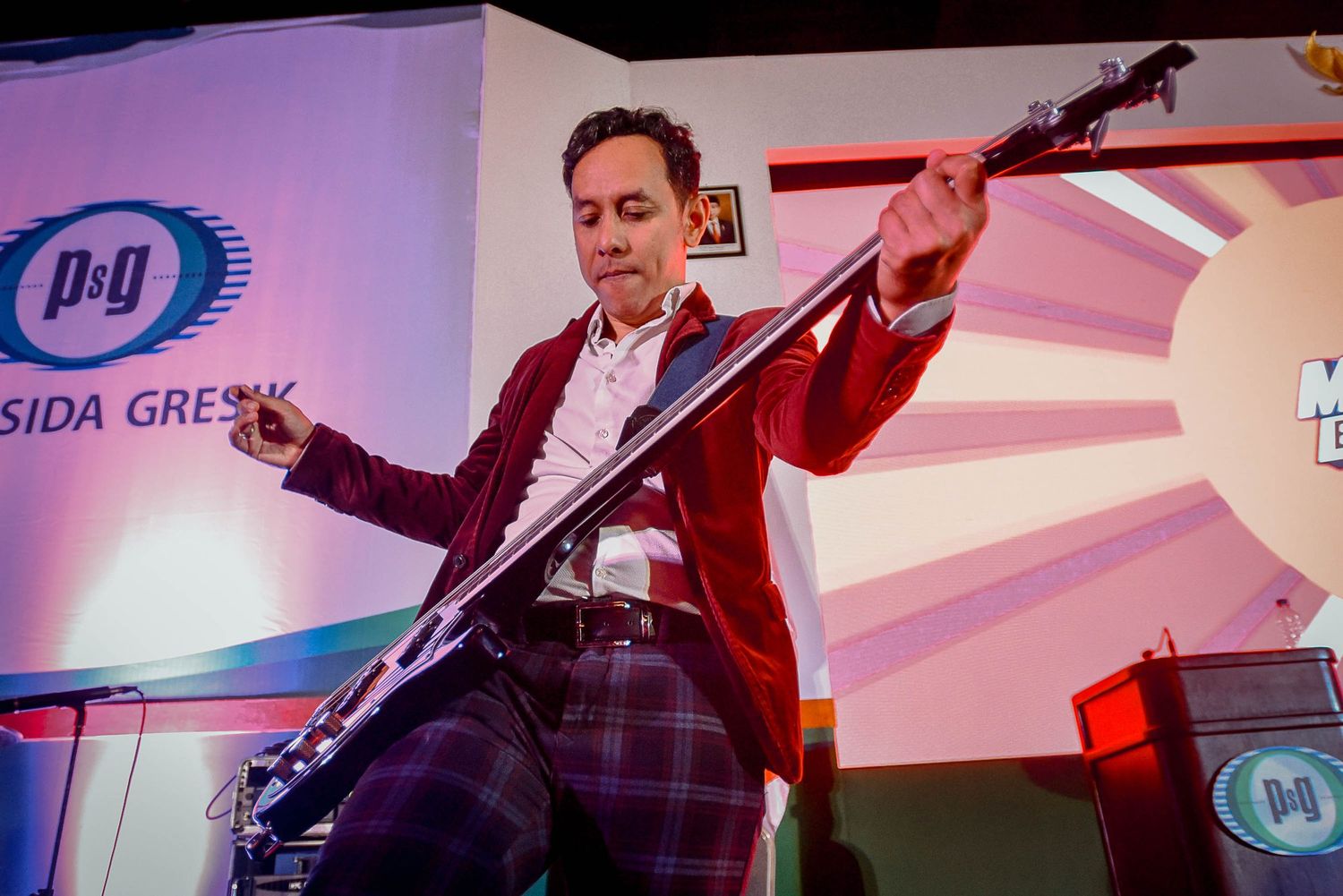 Musician plays bass guitar with enthusiasm on stage under red lighting.