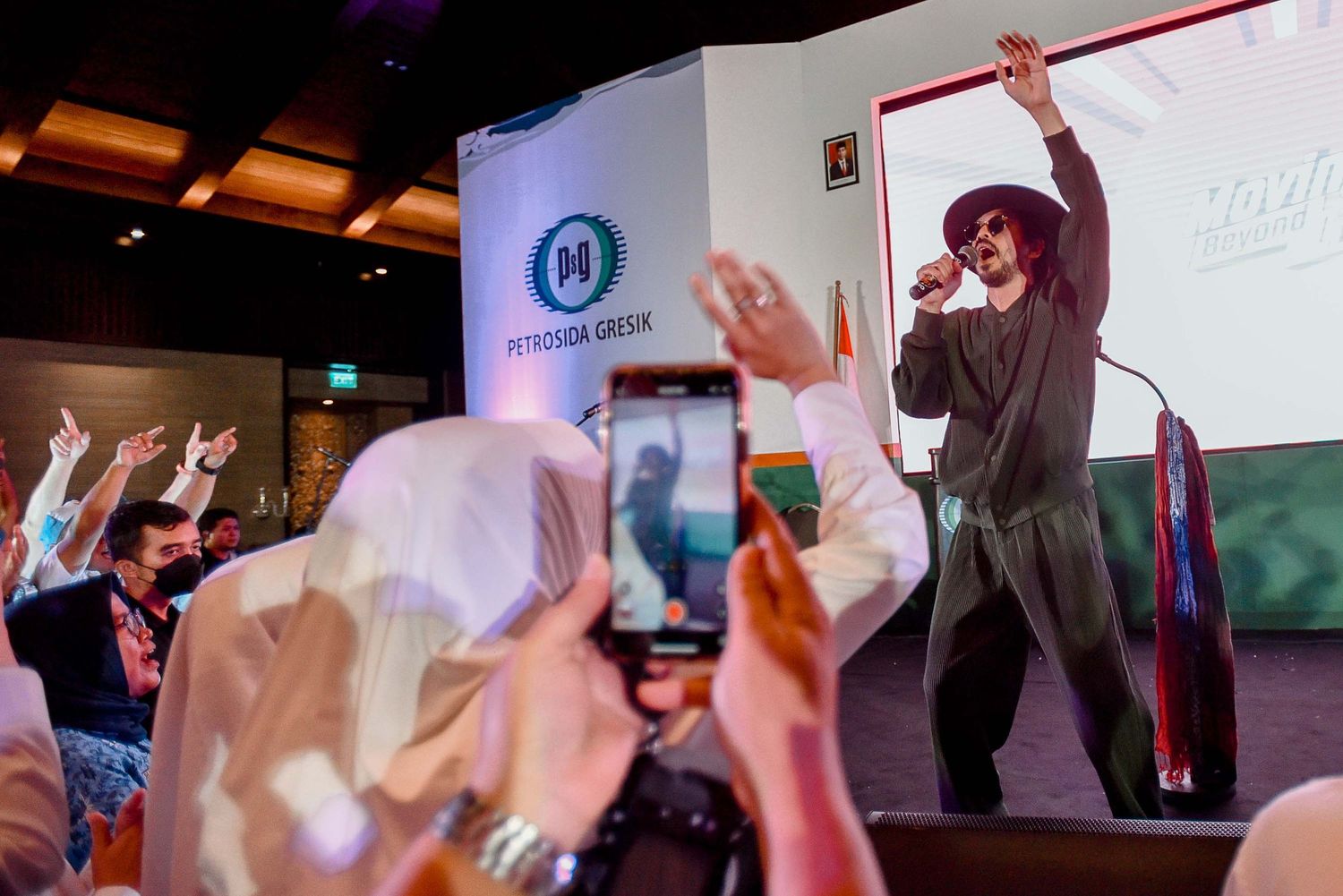 A performer in black raises their arms on stage as audience members hold up phones to capture the moment.
