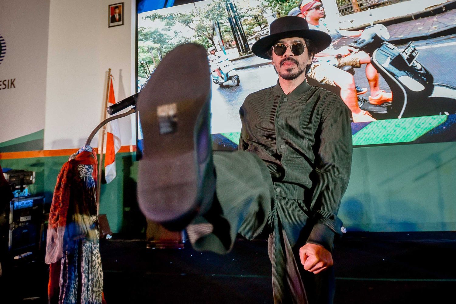 Performer in black hat reclines casually on stage with foot extended.