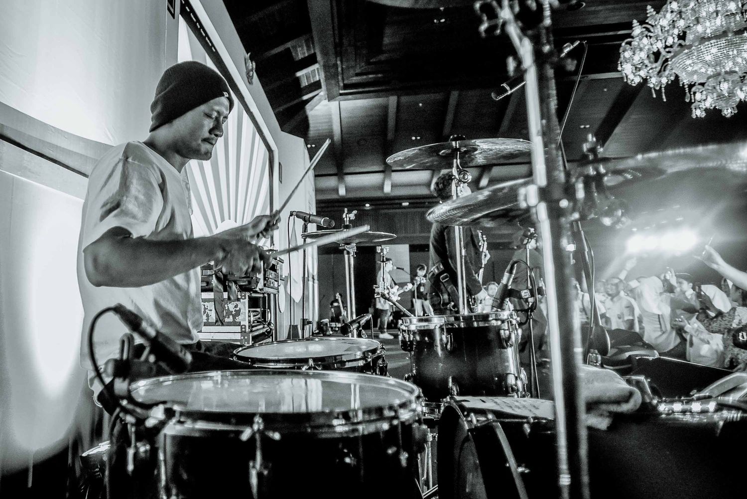 Black and white close-up shot of drummer performing behind drum kit on stage.