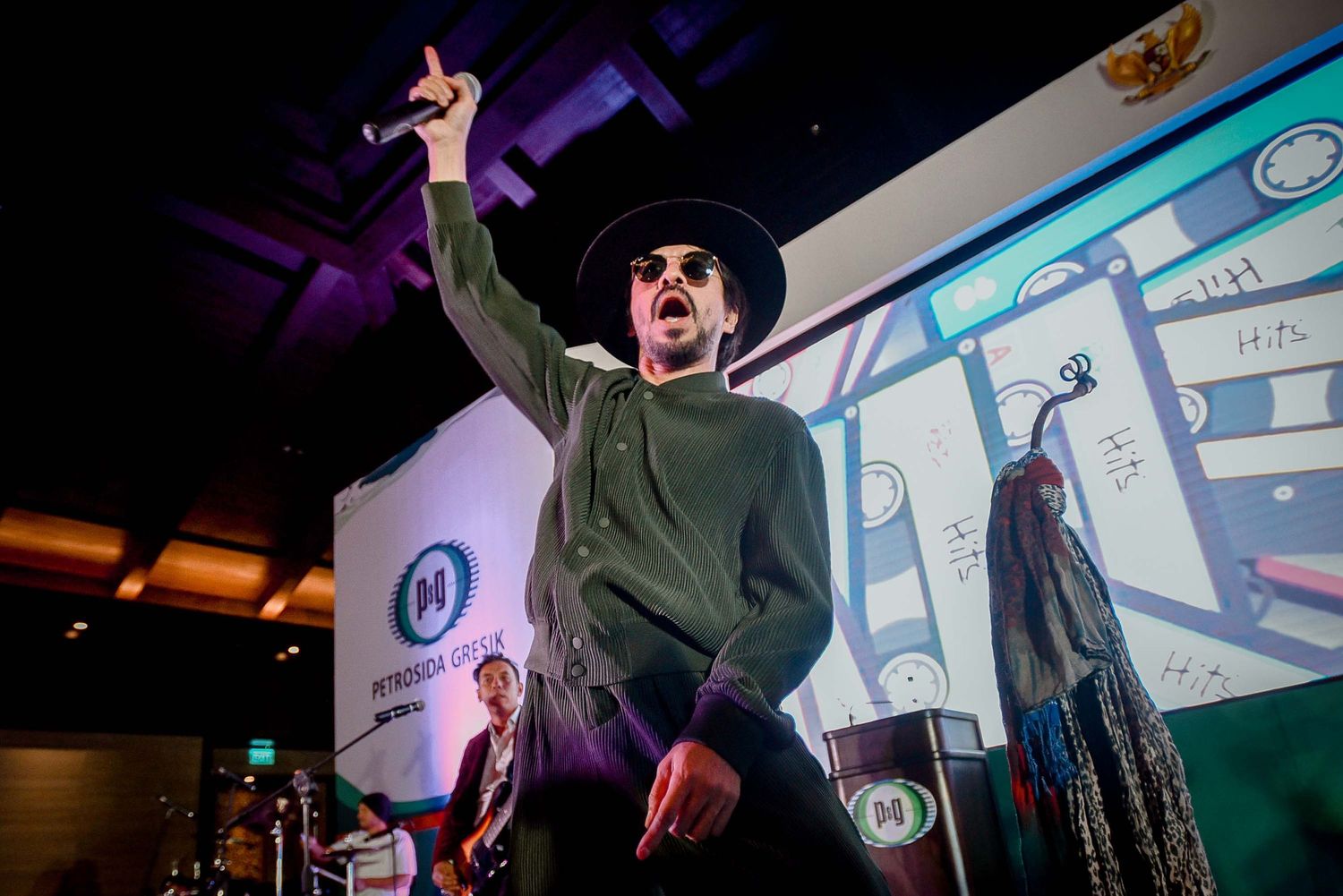 An energetic performer in a hat gestures dramatically on stage under purple lighting.
