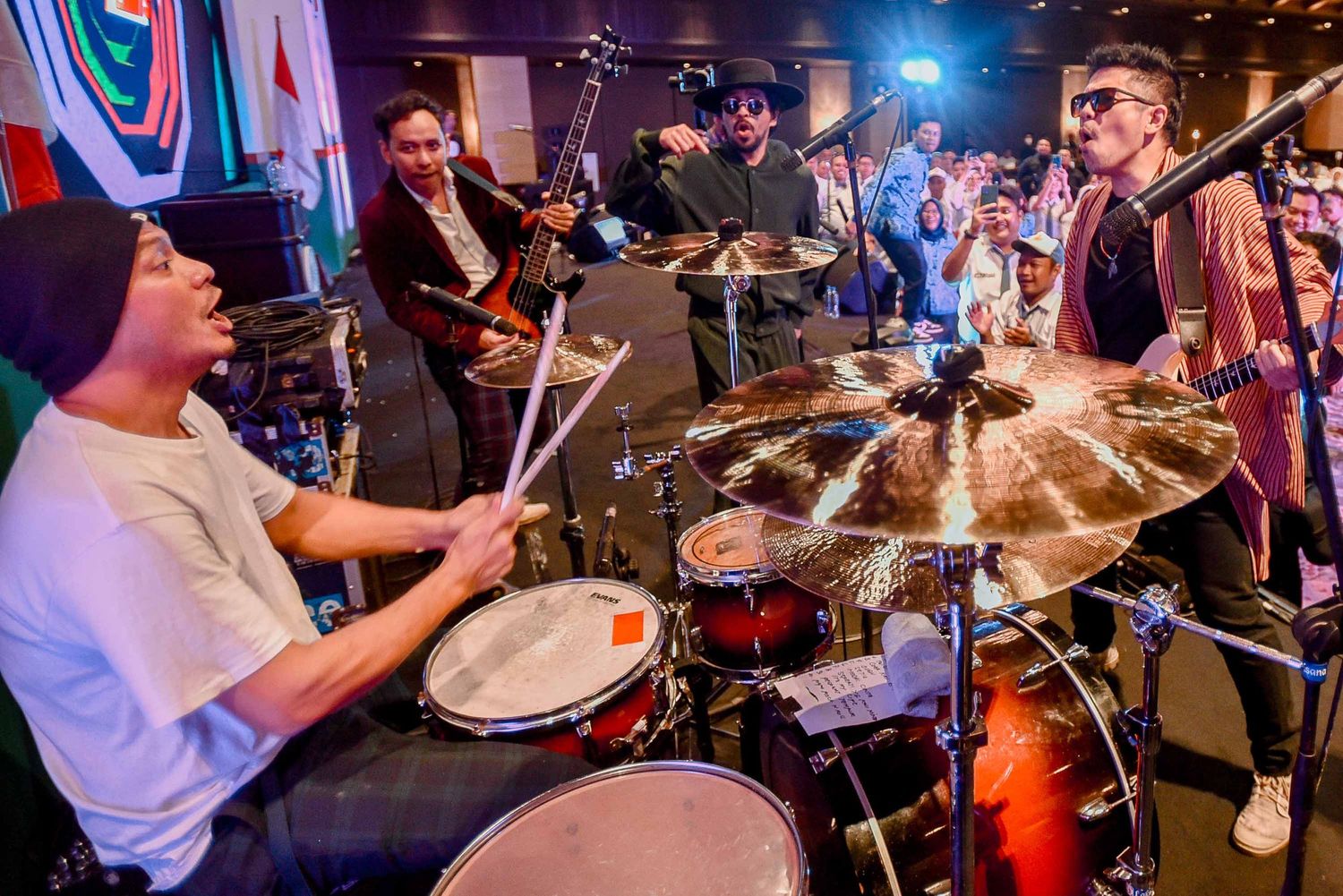 Drummer plays on stage with band members performing in colorful concert lighting.