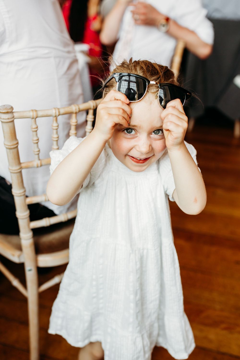 A playful child wearing sunglasses and a white dress at a wedding reception.