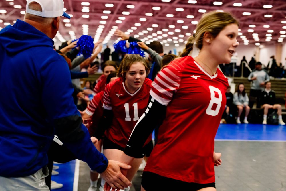 Young volleyball players in red uniforms high-fiving coaches during a tournament entrance.