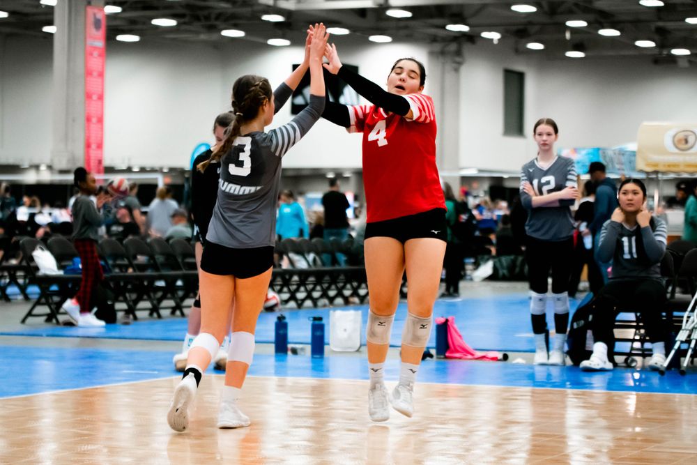 Two female volleyball players in red and gray uniforms high-fiving during an indoor match.