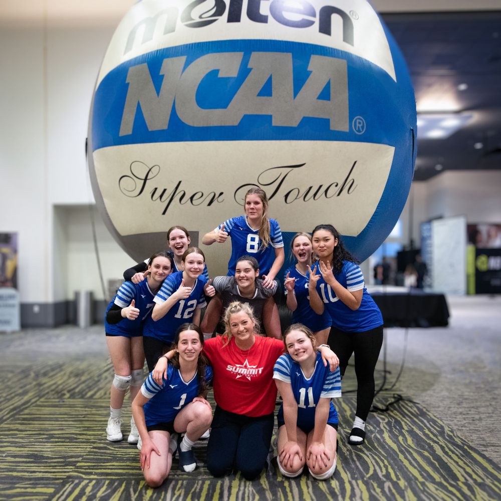Girls volleyball team poses in front of giant NCAA Molten Super Touch volleyball at tournament venue.