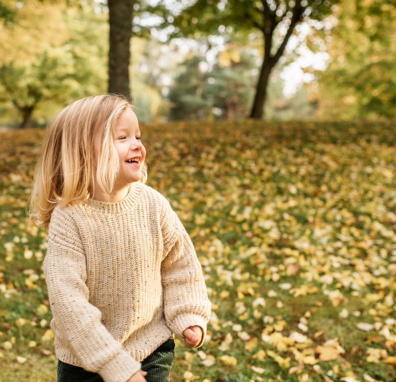 Young child laughing joyfully while wearing a cream knit sweater in an autumn setting.
