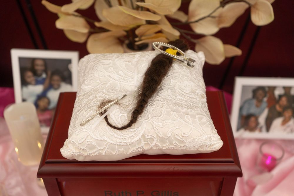 A decorative white lace pillow on a wooden box displaying a hair braid keepsake with family photos in background.