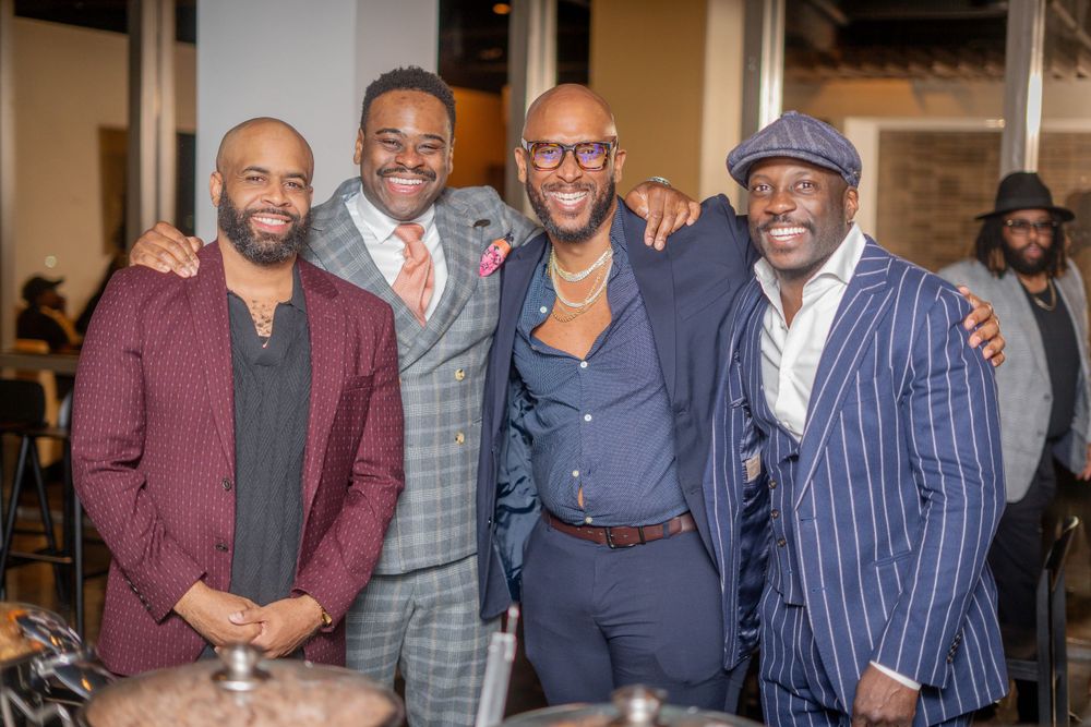 Four well-dressed men in suits smiling and posing together at a formal event.