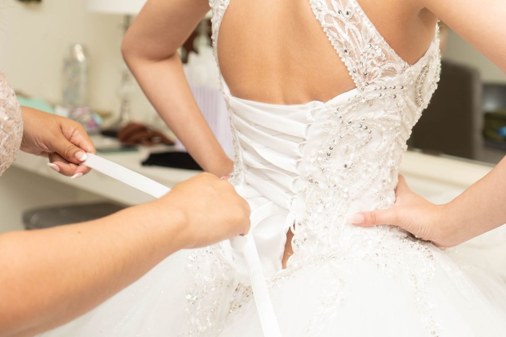 Bride in white lace corset wedding dress being laced up by assistant in bright dressing room.