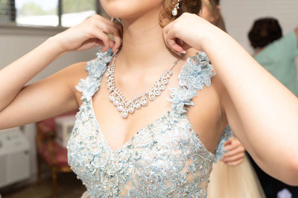 Woman in light blue floral embroidered gown adjusting a sparkling diamond necklace before a formal event.
