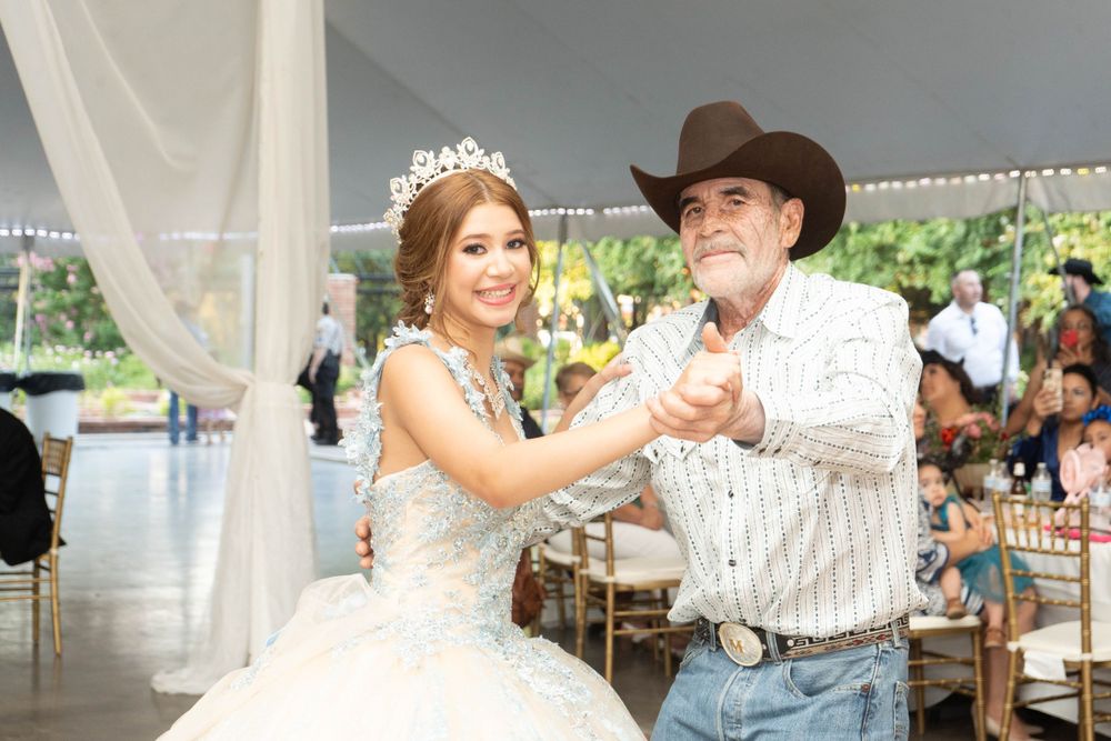 Young girl in blue quinceañera ball gown wearing tiara dances with older man in cowboy hat at reception.